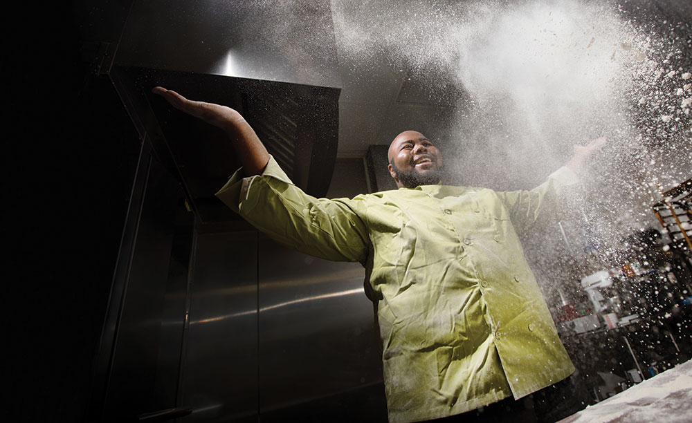 Jerron Corley, wearing a chef uniform, tosses flour in the air with a smile.